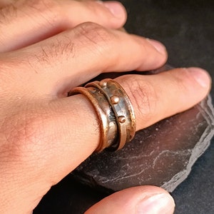 Puede incluir: Un anillo ancho de cobre y plata con textura martillada y pequeños acentos redondos. El anillo se lleva en un dedo.