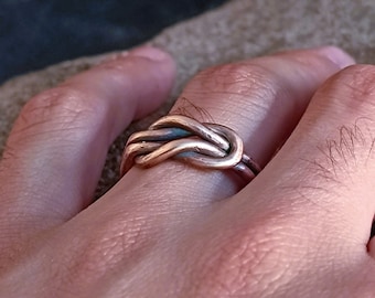 May include: A silver ring with a knot design. The ring is worn on a person's finger.