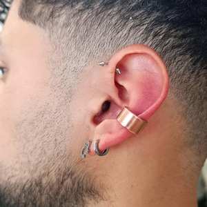 Puede incluir: Primer plano de una oreja con múltiples piercings. Un brazalete de oreja de color cobre está en la parte superior de la oreja, con un pendiente de aro plateado y un pequeño pendiente plateado en el lóbulo. Dos piercings plateados son visibles en la parte superior de la oreja.