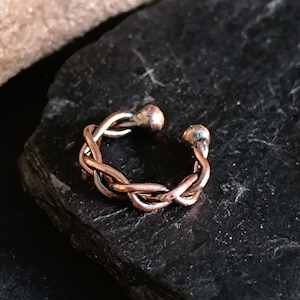 Könnte beinhalten: Ein geflochtener Ohrring aus Kupfer und Silber. Der Ohrring besteht aus zwei ineinander verschlungenen Drähten, einem aus Kupfer und einem aus Silber. Die Enden des Ohrrings sind abgerundet.