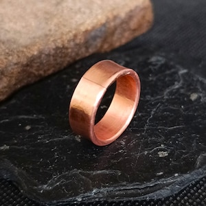 May include: A copper ring with a smooth, polished surface. The ring has a simple, band-style design and is displayed on a dark, textured surface. The ring's warm, reddish-brown color is highlighted by the light.
