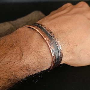 May include: A copper cuff bracelet with a hammered texture and a dark, oxidized stripe. The bracelet is worn on a wrist, showcasing its rustic, handcrafted design. The metal has a warm, reddish-brown hue.