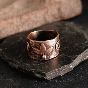 Op de afbeelding: Een koperen ring met een gehamerde textuur en een bloemmotief. De ring ligt op een donkergrijze stenen ondergrond.