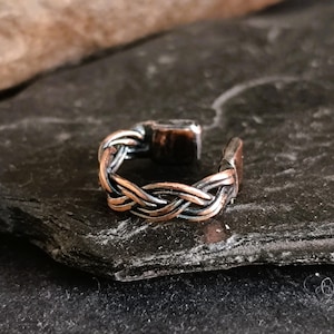 May include: A braided copper and silver ring with a hammered finish. The ring is open and has a slightly curved shape.