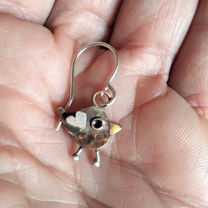 Op de afbeelding: Een handgemaakte oorbel in de vorm van een vogel met een zilveren haak. De vogel heeft een koperen lichaam, een zilveren kop met een hartdetail en een gele snavel. De oorbel heeft kleine zilveren pootjes en een zwart oog.