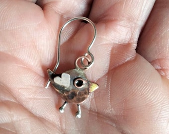 May include: A handcrafted bird-shaped earring with a silver hook. The bird features a copper body, a silver head with a heart detail, and a yellow beak. The earring has small silver legs and a black eye.