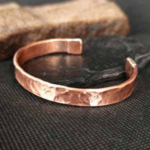 May include: A copper cuff bracelet with a hammered texture. The open-ended bracelet is a warm reddish-brown color. The cuff is resting on a black surface with a brown stone in the background.