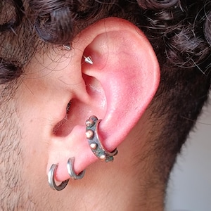 May include: Close-up of an ear with multiple piercings. A silver hoop earring and two smaller silver hoop earrings are visible. A cartilage piercing features a silver spike. The ear has a reddish hue.