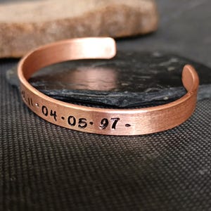 Puede incluir: Una pulsera de puño de cobre con texto estampado que dice "11-04-05-97 -". La pulsera tiene un diseño sencillo y abierto y un acabado pulido. La pulsera se muestra sobre una superficie oscura.