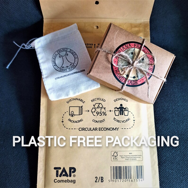 May include: A brown cardboard box with a red and white sticker that says "Adriana Franco Joyeria Hand Made" and a brown string bow. The box is sitting on a brown paper envelope with the text "Plastic Free Packaging" and a circular diagram showing a sustainable packaging process. The envelope also has the text "TAP Comebag 2/B" and a barcode.