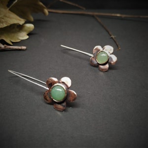 Pu&ograve; includere: Un paio di orecchini a forma di fiore. Ogni orecchino presenta un fiore color rame con un centro di pietra preziosa verde chiaro e un perno in filo d'argento. Gli orecchini sono su uno sfondo scuro.