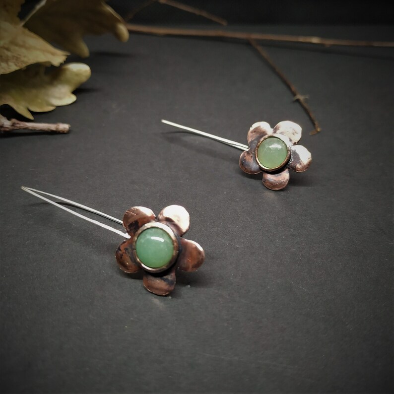 Pu&ograve; includere: Un paio di orecchini a forma di fiore. Ogni orecchino presenta un fiore color rame con un centro di pietra preziosa verde chiaro e un perno in filo d'argento. Gli orecchini sono su uno sfondo scuro.