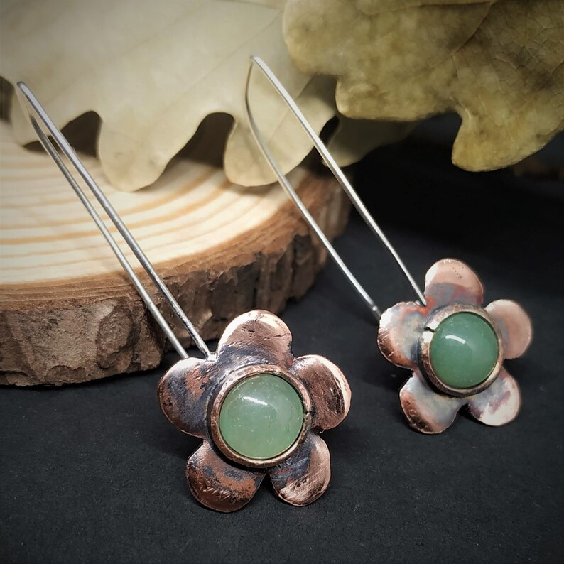 Pu&ograve; includere: Un paio di orecchini a forma di fiore. Ogni orecchino presenta un fiore a cinque petali color rame con una pietra rotonda verde chiaro al centro. Gli orecchini hanno lunghi fili color argento per l'orecchio. Gli orecchini sono esposti su una superficie scura.