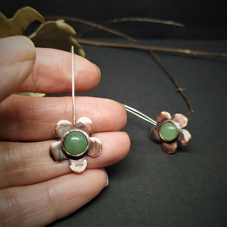 Pu&ograve; includere: Un paio di orecchini a forma di fiore fatti a mano. Ogni orecchino presenta un fiore di rame con un centro di pietra verde. Gli orecchini sono su un filo d'argento. Sfondo scuro.