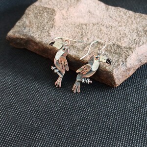 May include: A pair of toucan earrings made of copper and silver. The toucans are perched on a branch and have black eyes, orange beaks, and brown and white feathers.