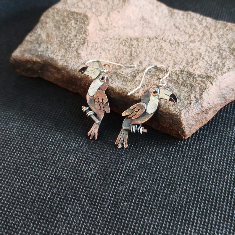 May include: A pair of toucan earrings made of copper and silver. The toucans are perched on a branch and have black eyes, orange beaks, and brown and white feathers.