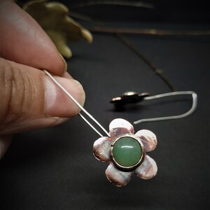 Pu&ograve; includere: Orecchini a forma di fiore con una gemma verde al centro. Gli orecchini hanno un design a fiore color rame e un gancio in filo d'argento. La gemma &egrave; di colore verde chiaro. Gli orecchini sono mostrati su uno sfondo scuro.