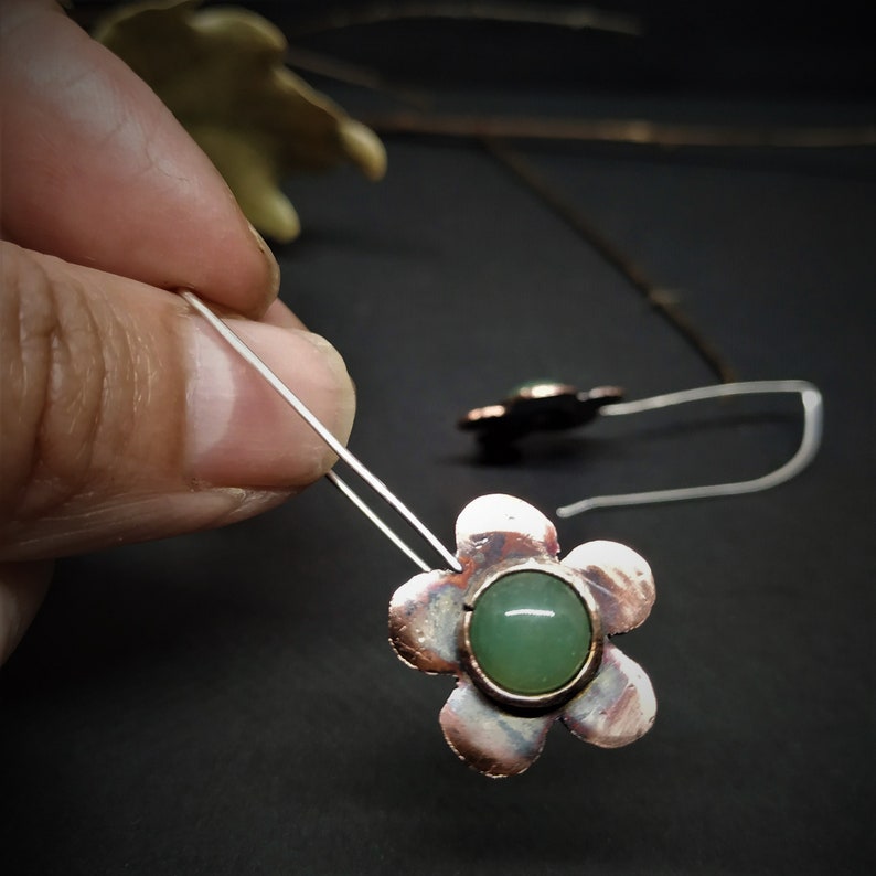Pu&ograve; includere: Orecchini a forma di fiore con una gemma verde al centro. Gli orecchini hanno un design a fiore color rame e un gancio in filo d'argento. La gemma &egrave; di colore verde chiaro. Gli orecchini sono mostrati su uno sfondo scuro.