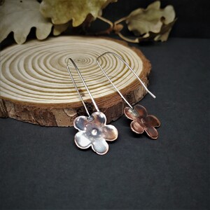 Pu&ograve; includere: Un paio di orecchini fatti a mano con un design a forma di fiore. Gli orecchini hanno un ciondolo a forma di fiore color rame con un gancio color argento. Gli orecchini sono esposti su una superficie di legno con uno sfondo scuro.