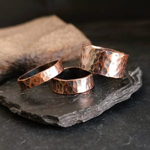 May include: Three hammered copper rings of varying widths, stacked on a dark gray stone surface.