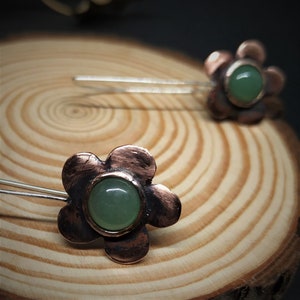 Pu&ograve; includere: Primo piano di due forcine per capelli a forma di fiore. Ogni forcina presenta un fiore in metallo color rame con un centro di pietra verde. Le forcine sono poste su uno sfondo di legno, evidenziando i dettagli artigianali e i toni naturali.