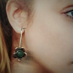 Pu&ograve; includere: Un singolo orecchino d'argento con un ciondolo a forma di fiore. Il ciondolo ha un fiore color rame con un centro di pietra verde. L'orecchino &egrave; mostrato su uno sfondo sfocato.