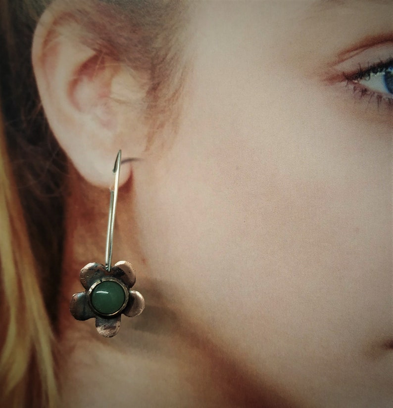 Pu&ograve; includere: Un singolo orecchino d'argento con un ciondolo a forma di fiore. Il ciondolo ha un fiore color rame con un centro di pietra verde. L'orecchino &egrave; mostrato su uno sfondo sfocato.