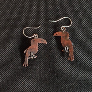 May include: A pair of copper toucan earrings with silver hooks. The toucans are facing opposite directions.