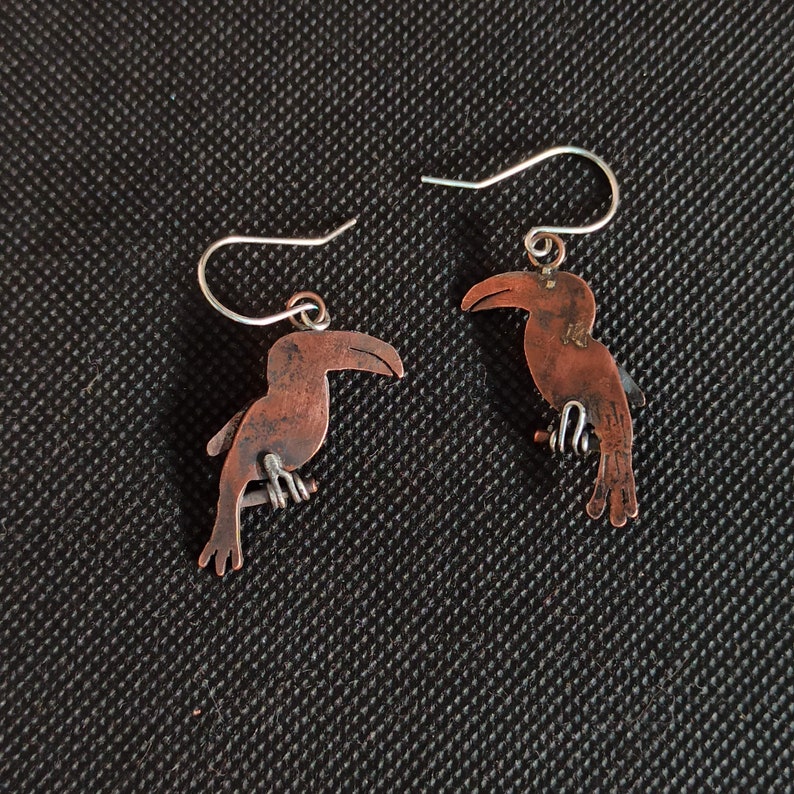 May include: A pair of copper toucan earrings with silver hooks. The toucans are facing opposite directions.