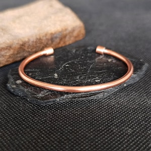 Op de afbeelding: Een koperkleurige manchetarmband met afgeronde uiteinden. De armband wordt gepresenteerd op een donkere, getextureerde ondergrond, met een ruwe, bruine steen op de achtergrond. De armband heeft een eenvoudig, minimalistisch ontwerp.