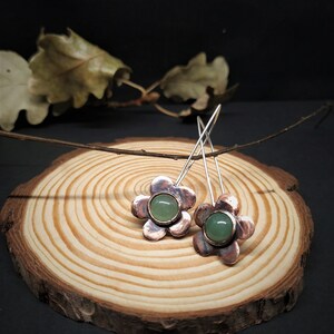 Pu&ograve; includere: Orecchini a forma di fiore con metallo color rame e un centro in pietra verde. Gli orecchini sono esposti su una fetta di legno su sfondo scuro. Gli orecchini hanno lunghi ganci color argento.