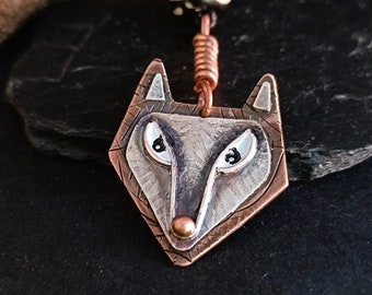 May include: A silver and copper wolf pendant with a textured surface and black eyes. The pendant is attached to a copper chain.