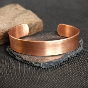 Può includere: Un bracciale a polsino in rame con finitura spazzolata. Il bracciale aperto è esposto su una pietra grigio scuro, con una roccia dalla consistenza ruvida sullo sfondo. Il bracciale è largo circa 2 cm.