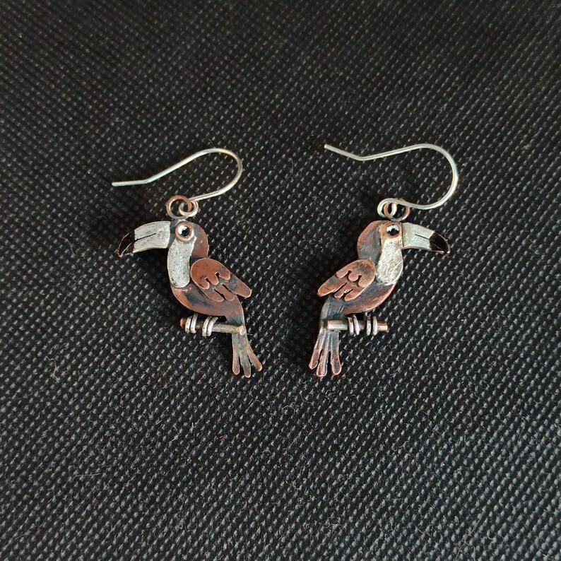 May include: A pair of toucan earrings made of copper and silver. The toucans are perched on a branch and have black, white, and brown details.