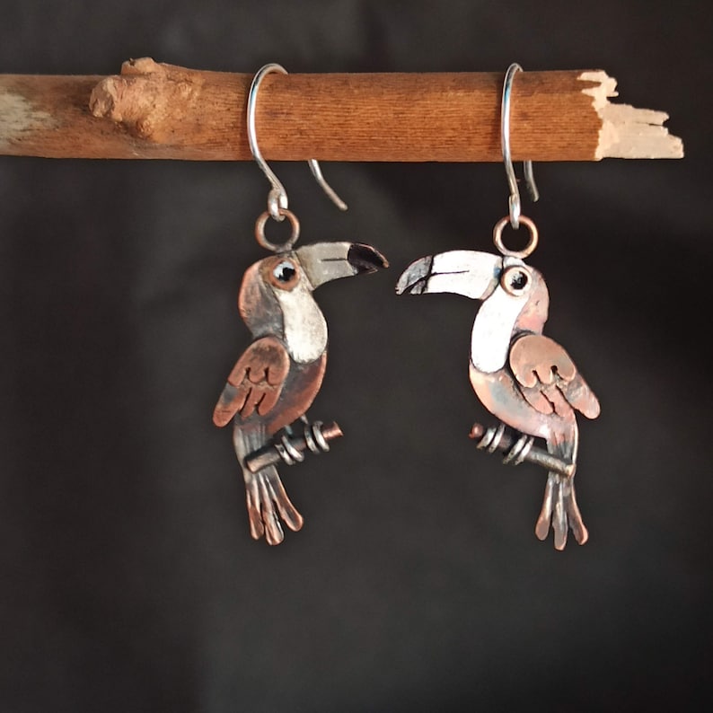 May include: A pair of toucan earrings made of copper and silver. The toucans are painted with white, brown, and black accents. The earrings are hanging from silver hooks.