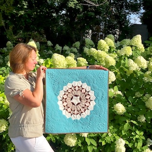 May include: A woman holds a blue quilted wall hanging with a brown and white geometric design. The design is a circular pattern with a starburst shape in the center. The wall hanging is surrounded by a brown border.