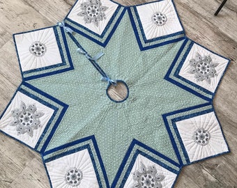 Puede incluir: Una falda de árbol de Navidad en forma de estrella, en tonos azul claro y blanco. Presenta una abertura central y está adornada con diseños florales bordados y lazos azules. El diseño incluye un patrón acolchado y bordes azul oscuro.