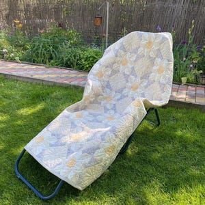 Op de afbeelding: Een bloemmotief quilt in crème, geel en lavendel is over een blauwe metalen klapstoel gedrapeerd. Het ontwerp van de quilt heeft herhalende bloemvormen. De stoel staat op een groen gazon, met een tuin zichtbaar op de achtergrond.
