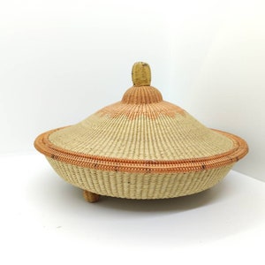 May include: A woven basket with a lid, featuring a natural beige colour with orange accents. The basket has a round base with three legs and a domed lid with a small knob on top.
