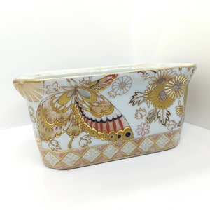 Könnte beinhalten: Ein weißer Keramik-Pflanztopf mit goldfarbenem Rand und einem aufwendigen Blumen- und Schmetterlingsdesign. Der Topf hat eine rechteckige Form mit abgerundeten Kanten.