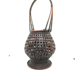 May include: A dark brown woven bamboo basket with a round base and a single handle. The basket has a lattice pattern and is ideal for displaying flowers or other decorative items.