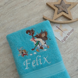 Peut inclure: Une serviette de bain turquoise vif ornée d'une broderie représentant les personnages de dessin animé Astérix et Obélix en train de courir, avec le prénom Felix brodé en lettres cursives blanches et une bordure texturée en bas.