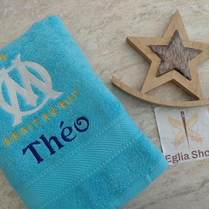 Peut inclure: Une serviette turquoise vif avec le logo blanc brodé de l'Olympique de Marseille, le texte jaune DROIT AU BUT et le prénom Théo en bleu foncé. À côté se trouvent une étoile décorative en bois clair et une étiquette blanche Egli Shop.