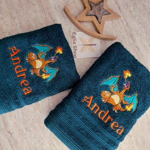 Puede incluir: Dos toallas de baño de color verde azulado oscuro con el texto naranja bordado Andrea y una criatura colorida similar a un dragón que escupe fuego. Las toallas tienen una superficie texturizada con bandas acanaladas horizontales.