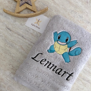 Peut inclure: Une serviette gris clair avec une broderie représentant une tortue bleue et jaune et le prénom Lennart en écriture noire. Un ornement en bois en forme d'étoile et une petite carte blanche avec le texte Eglia Shop sont placés à côté.