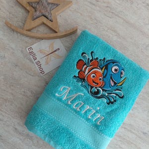 Peut inclure: Une serviette de bain turquoise vif avec une broderie représentant deux poissons-clowns et un poisson chirurgien nageant dans l'eau. Le prénom Marin est brodé en lettres cursives blanches sous les poissons. Un ornement en étoile en bois est présent.