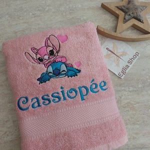 Può includere: Un morbido asciugamano da bagno rosa con un ricamo che raffigura il personaggio blu Stitch e il personaggio rosa Angel con due piccoli cuori rosa. Il nome Cassiopée è ricamato in blu in corsivo sotto i personaggi sull'asciugamano.