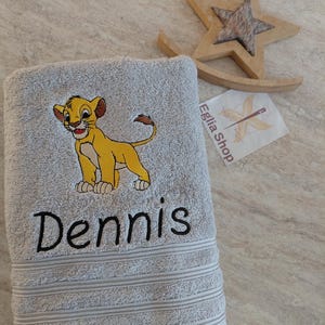 Peut inclure: Une serviette de bain gris clair avec une broderie représentant un lionceau jaune et le prénom Dennis en fil noir. La serviette présente une texture côtelée sur le bord inférieur. Un ornement en bois en forme d'étoile et une étiquette blanche avec le texte Eglia Shop sont visibles à proximité.