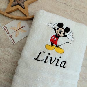 Peut inclure: Une serviette en éponge blanche avec une broderie représentant Mickey Mouse en short rouge et chaussures jaunes, accompagnée du prénom Livia cousu en lettres cursives noires sous le personnage. La serviette présente trois bandes texturées.