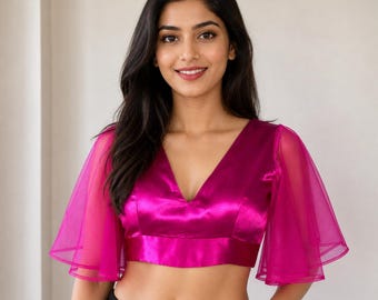 Puede incluir: Un top corto de color magenta con cuello en V profundo y mangas transparentes acampanadas. La parte superior está hecha de un material brillante, similar al satén. El modelo lleva pantalones negros.